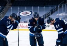 Winnipeg Jets players Mark Scheifele (#55), Kyle Connor (#81), and Josh Morrissey (#44) stand dejected on the ice with their heads bowed in frustration inside an empty arena.