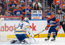 An Edmonton Oilers player with the puck skates in on Buffalo Sabres goaltender Alex Lyon, who is positioned in his crease. Behind the glass in the crowd, a female fan in an Oilers jersey holds a sign that reads "COME TO EDMONTON ALEX!".