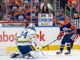 An Edmonton Oilers player with the puck skates in on Buffalo Sabres goaltender Alex Lyon, who is positioned in his crease. Behind the glass in the crowd, a female fan in an Oilers jersey holds a sign that reads "COME TO EDMONTON ALEX!".