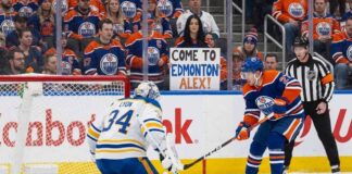 An Edmonton Oilers player with the puck skates in on Buffalo Sabres goaltender Alex Lyon, who is positioned in his crease. Behind the glass in the crowd, a female fan in an Oilers jersey holds a sign that reads "COME TO EDMONTON ALEX!".