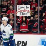 Vancouver Canucks forward Elias Pettersson (40) skates on the ice during a game. Behind the glass, a female fan in a Carolina Hurricanes jersey holds up a sign that reads, "Come to Carolina Elias!", while surrounded by other cheering Hurricanes fans.