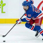 NY Rangers Trade Rumors: Is Erik Gustafsson the Adam Fox Injury Fix? New York Rangers defenseman Erik Gustafsson, wearing number 56 in a blue home uniform and blue helmet, skates along the boards handling the puck, with GFL and Coca-Cola advertisements visible in the background.