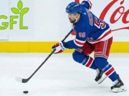 NY Rangers Trade Rumors: Is Erik Gustafsson the Adam Fox Injury Fix? New York Rangers defenseman Erik Gustafsson, wearing number 56 in a blue home uniform and blue helmet, skates along the boards handling the puck, with GFL and Coca-Cola advertisements visible in the background.