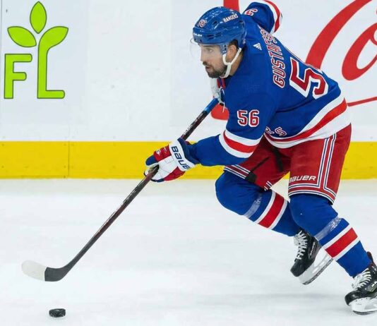 NY Rangers Trade Rumors: Is Erik Gustafsson the Adam Fox Injury Fix? New York Rangers defenseman Erik Gustafsson, wearing number 56 in a blue home uniform and blue helmet, skates along the boards handling the puck, with GFL and Coca-Cola advertisements visible in the background.