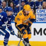 Toronto Maple Leafs captain Auston Matthews battles Nashville Predators forward Jonathan Marchessault for the puck along the boards. A Leafs fan in the background holds up a handmade sign that reads, "COME TO TORONTO JONATHAN!" amidst ongoing trade rumors.