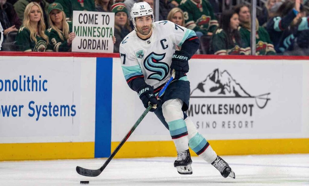 Seattle Kraken captain Jordan Eberle, wearing his white away #7 jersey, skates with the puck on the ice during a game. In the background, fans wearing Minnesota Wild jerseys watch from the stands, including one woman in the front row holding up a white sign with black text that reads, "COME TO MINNESOTA JORDAN!".
