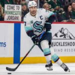 Seattle Kraken captain Jordan Eberle, wearing his white away #7 jersey, skates with the puck on the ice during a game. In the background, fans wearing Minnesota Wild jerseys watch from the stands, including one woman in the front row holding up a white sign with black text that reads, "COME TO MINNESOTA JORDAN!".