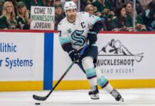 Seattle Kraken captain Jordan Eberle, wearing his white away #7 jersey, skates with the puck on the ice during a game. In the background, fans wearing Minnesota Wild jerseys watch from the stands, including one woman in the front row holding up a white sign with black text that reads, "COME TO MINNESOTA JORDAN!".
