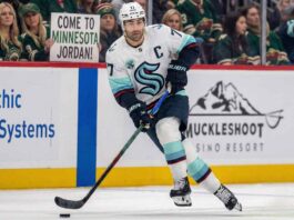 Seattle Kraken captain Jordan Eberle, wearing his white away #7 jersey, skates with the puck on the ice during a game. In the background, fans wearing Minnesota Wild jerseys watch from the stands, including one woman in the front row holding up a white sign with black text that reads, "COME TO MINNESOTA JORDAN!".