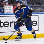 St. Louis Blues defenseman Justin Faulk (#72) skates with the puck on the ice, looking towards the stands where a female fan holds a handwritten cardboard sign that reads "We will miss you Justin!" behind the protective glass.