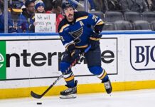 St. Louis Blues defenseman Justin Faulk (#72) skates with the puck on the ice, looking towards the stands where a female fan holds a handwritten cardboard sign that reads "We will miss you Justin!" behind the protective glass.