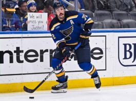 St. Louis Blues defenseman Justin Faulk (#72) skates with the puck on the ice, looking towards the stands where a female fan holds a handwritten cardboard sign that reads "We will miss you Justin!" behind the protective glass.