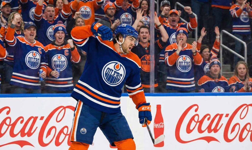Forward Kiefer Sherwood in a blue Oilers home jersey, yelling and pumping his fist in passionate celebration against the arena glass.