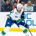 Vancouver Canucks winger Kiefer Sherwood #44 skates with the puck during an NHL game, while San Jose Sharks fans behind the plexiglass hold a sign reading "Come to San Jose Kiefer!".