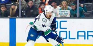 Vancouver Canucks winger Kiefer Sherwood #44 skates with the puck during an NHL game, while San Jose Sharks fans behind the plexiglass hold a sign reading "Come to San Jose Kiefer!".