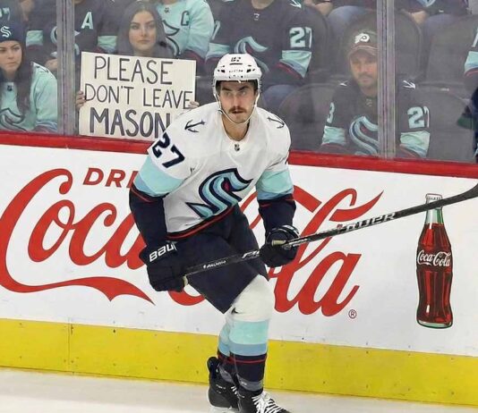 Seattle Kraken winger Mason Marchment (#27) stands on the ice near the boards during a game, wearing his white away jersey and helmet. He has a prominent mustache. Behind the protective glass, a female fan with long dark hair holds up a handwritten sign that reads, "PLEASE DON'T LEAVE MASON."