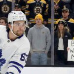 Rumor: Boston Bruins Linked to Toronto Maple Leafs Winger Matias Maccelli A photo of Toronto Maple Leafs forward Matias Maccelli, wearing his white #63 away jersey, skating in the foreground. Behind the rink glass, a crowd of Boston Bruins fans watches, some in team jerseys and others in casual clothes like hoodies and jackets. A woman in a Bruins jersey holds a handwritten white sign that reads "BOSTON WANTS YOU MATIAS!".