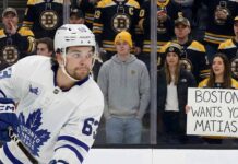 A photo of Toronto Maple Leafs forward Matias Maccelli, wearing his white #63 away jersey, skating in the foreground. Behind the rink glass, a crowd of Boston Bruins fans watches, some in team jerseys and others in casual clothes like hoodies and jackets. A woman in a Bruins jersey holds a handwritten white sign that reads "BOSTON WANTS YOU MATIAS!".