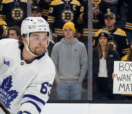 Rumor: Boston Bruins Linked to Toronto Maple Leafs Winger Matias Maccelli A photo of Toronto Maple Leafs forward Matias Maccelli, wearing his white #63 away jersey, skating in the foreground. Behind the rink glass, a crowd of Boston Bruins fans watches, some in team jerseys and others in casual clothes like hoodies and jackets. A woman in a Bruins jersey holds a handwritten white sign that reads "BOSTON WANTS YOU MATIAS!".
