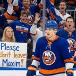 New York Islanders player Maxim Tsyplakov stands on the ice near the glass, looking towards cheering fans. A blonde woman in the front row holds a handwritten sign that reads, "Please don't leave Maxim!".