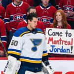 Montreal Canadiens Goalie Nightmare: Why a Trade Feels Inevitable St. Louis Blues goaltender Jordan Binnington, wearing his white number 50 jersey, stands on the ice looking downward. Behind the rink glass, a crowd of fans wearing red Montreal Canadiens jerseys looks on. In the front row, a smiling woman with red hair holds up a white handwritten sign that reads, "Come to Montreal Jordan!".