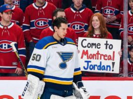Montreal Canadiens Goalie Nightmare: Why a Trade Feels Inevitable St. Louis Blues goaltender Jordan Binnington, wearing his white number 50 jersey, stands on the ice looking downward. Behind the rink glass, a crowd of fans wearing red Montreal Canadiens jerseys looks on. In the front row, a smiling woman with red hair holds up a white handwritten sign that reads, "Come to Montreal Jordan!".