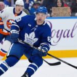 Toronto Maple Leafs Mattias Maccelli in a blue jersey, number 63, skates with the puck during a hockey game. Behind him, a New York Islanders player in a white jersey, number 27, follows. In the background stands, a fan holds up a cardboard sign that reads "BYE-BYE MATTIAS!".