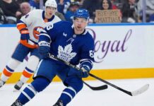 Toronto Maple Leafs Mattias Maccelli in a blue jersey, number 63, skates with the puck during a hockey game. Behind him, a New York Islanders player in a white jersey, number 27, follows. In the background stands, a fan holds up a cardboard sign that reads "BYE-BYE MATTIAS!".