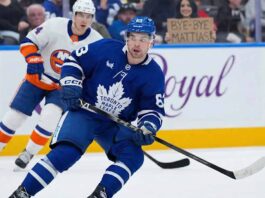 Toronto Maple Leafs Mattias Maccelli in a blue jersey, number 63, skates with the puck during a hockey game. Behind him, a New York Islanders player in a white jersey, number 27, follows. In the background stands, a fan holds up a cardboard sign that reads "BYE-BYE MATTIAS!".