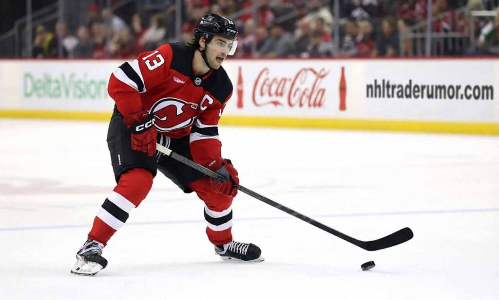 New Jersey Devils captain Nico Hischier (#13) in a low skating stance on the ice, looking forward with his stick near a puck.