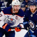 Edmonton Oilers captain Connor McDavid (#97) in a white away jersey yelling while skating intensely alongside Winnipeg Jets forward Kyle Connor (#81) in a dark blue home jersey during an NHL game.