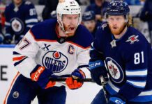Edmonton Oilers captain Connor McDavid (#97) in a white away jersey yelling while skating intensely alongside Winnipeg Jets forward Kyle Connor (#81) in a dark blue home jersey during an NHL game.