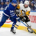 Toronto Maple Leafs captain Auston Matthews (left, blue home jersey) and Pittsburgh Penguins captain Sidney Crosby (right, white away jersey) battle fiercely with sticks crossed for the puck along the rink boards, with a crowd of Maple Leafs fans visible behind the glass.