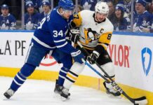 Toronto Maple Leafs captain Auston Matthews (left, blue home jersey) and Pittsburgh Penguins captain Sidney Crosby (right, white away jersey) battle fiercely with sticks crossed for the puck along the rink boards, with a crowd of Maple Leafs fans visible behind the glass.