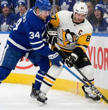 Toronto Maple Leafs captain Auston Matthews (left, blue home jersey) and Pittsburgh Penguins captain Sidney Crosby (right, white away jersey) battle fiercely with sticks crossed for the puck along the rink boards, with a crowd of Maple Leafs fans visible behind the glass.