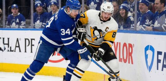 Toronto Maple Leafs captain Auston Matthews (left, blue home jersey) and Pittsburgh Penguins captain Sidney Crosby (right, white away jersey) battle fiercely with sticks crossed for the puck along the rink boards, with a crowd of Maple Leafs fans visible behind the glass.
