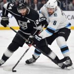 Los Angeles Kings center Phillip Danault (24), wearing a black home jersey, battles intensely for the puck along the ice with Utah forward Nick Schmaltz (8), wearing a white away jersey, during an NHL game.