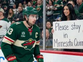 Quinn Hughes Trade: Why the Wild’s Massive Cup Gamble May Gift the Devils a Dynasty Minnesota Wild Quinn Hughes, wearing his green number 43 jersey, looks towards the crowd from the ice as a female fan holds up a white sign that reads "Please stay in Minnesota Quinn" during a game.