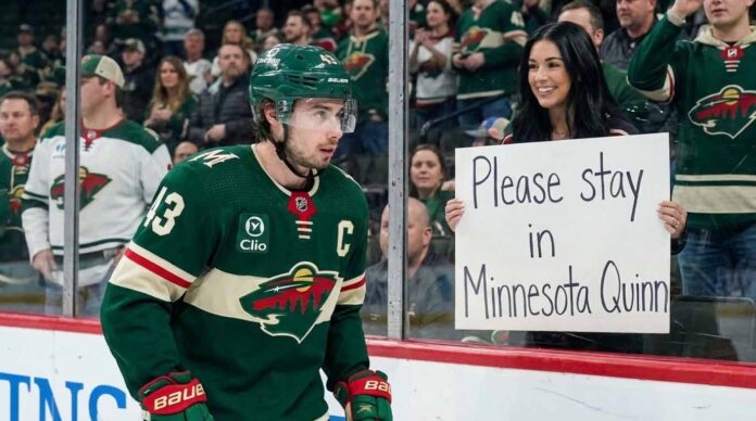 Minnesota Wild Quinn Hughes, wearing his green number 43 jersey, looks towards the crowd from the ice as a female fan holds up a white sign that reads "Please stay in Minnesota Quinn" during a game.