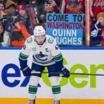 Why the Canucks Might Actually Trade Quinn Hughes to the Capitals Vancouver Canucks captain Quinn Hughes stands on the ice in his away jersey, looking towards the boards where a fan holds a sign that reads 'Come to Washington Quinn Hughes'.