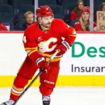 A photo of Calgary Flames defenseman Rasmus Andersson, wearing his red number 4 jersey, skating with the puck on the ice during a game. In the background, a blonde female fan holds a sign that reads, "Please don't leave Rasmus."