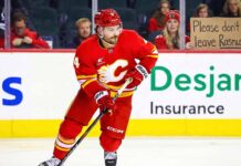 A photo of Calgary Flames defenseman Rasmus Andersson, wearing his red number 4 jersey, skating with the puck on the ice during a game. In the background, a blonde female fan holds a sign that reads, "Please don't leave Rasmus."