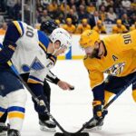 St. Louis Blues captain Brayden Schenn (#10) in a white away jersey and Nashville Predators alternate captain Ryan O'Reilly (#90) in a yellow home jersey crouch for a faceoff as a linesman drops the puck between their sticks during an NHL game.