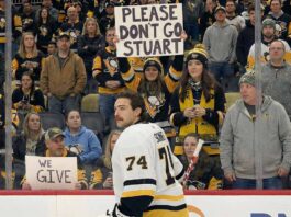 Stuart Skinner, wearing his white #74 Pittsburgh Penguins jersey, stands on the ice looking towards the fans behind the plexiglass. A fan in the center holds up a white sign with black letters that reads "PLEASE DON'T GO STUART".