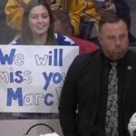 ormer Toronto Maple Leafs assistant coach Marc Savard stands on the bench looking serious while a fan behind the glass holds a handwritten sign reading We will miss you Marc.