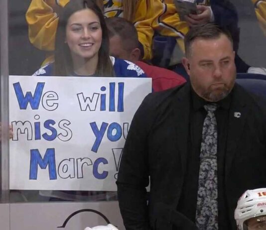 ormer Toronto Maple Leafs assistant coach Marc Savard stands on the bench looking serious while a fan behind the glass holds a handwritten sign reading We will miss you Marc.