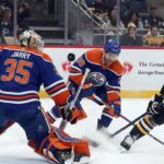Edmonton Oilers goaltender Tristan Jarry (#35), wearing a blue and orange uniform and matching pads, makes a save in the butterfly position during a game against the Pittsburgh Penguins, with an Oilers teammate and a Penguins player near the crease.