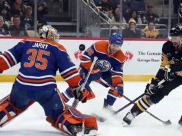 Edmonton Oilers goaltender Tristan Jarry (#35), wearing a blue and orange uniform and matching pads, makes a save in the butterfly position during a game against the Pittsburgh Penguins, with an Oilers teammate and a Penguins player near the crease.
