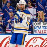Buffalo Sabres forward Alex Tuch (89) stands on the ice looking towards the crowd behind the glass, where a female fan holds a handwritten sign that reads "COME TO NEW YORK ALEX!" surrounded by New York Rangers supporters.