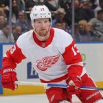 Detroit Red Wings forward Alexis Lafrenière, wearing number 13 in a white away jersey with red trim and the winged wheel logo, skates on the ice holding his stick during a game.